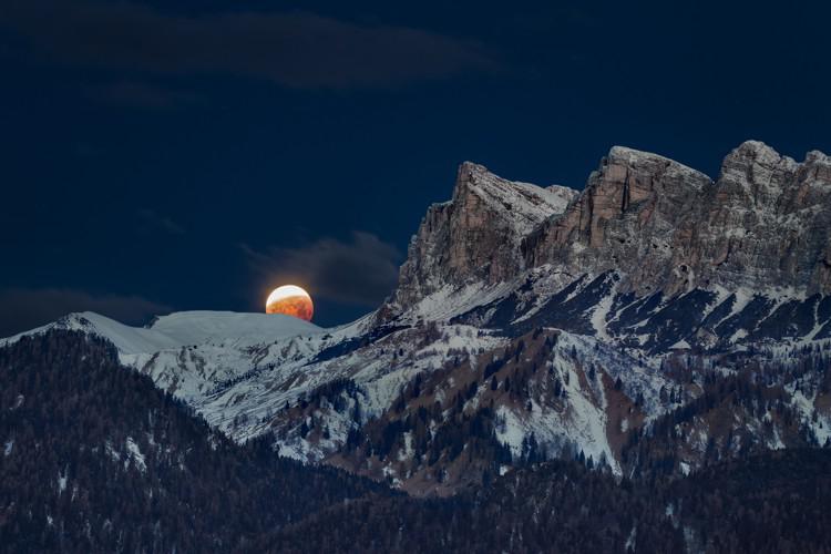 The Moon behind jagged, snowy mountains. The lower two thirds of the Moon are darker and redder than the upper third