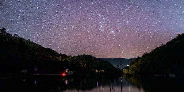 A starry sky is reflected in a lake in a valley. The streak of a meteor goes from the top right to the middle of the image