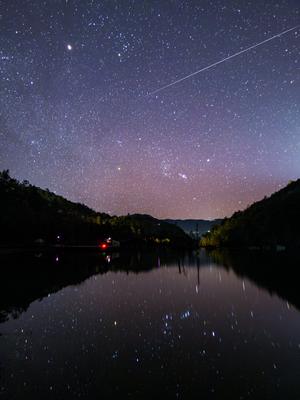 Un ciel étoilé se reflète dans un lac situé dans une vallée. La traînée d'un météore va du haut à droite au milieu de l'image.
