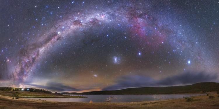 La curva della Via Lattea è tagliata a sinistra da un enorme arco scuro. In basso, due macchie luminose e diffuse.