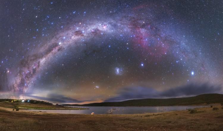 La curva della Via Lattea è tagliata a sinistra da un enorme arco scuro. In basso, due macchie luminose e diffuse.