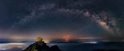 The Milky Way arches over a mountain-top building. Its diffuse glow is broken by dark patches and is brighter on the right