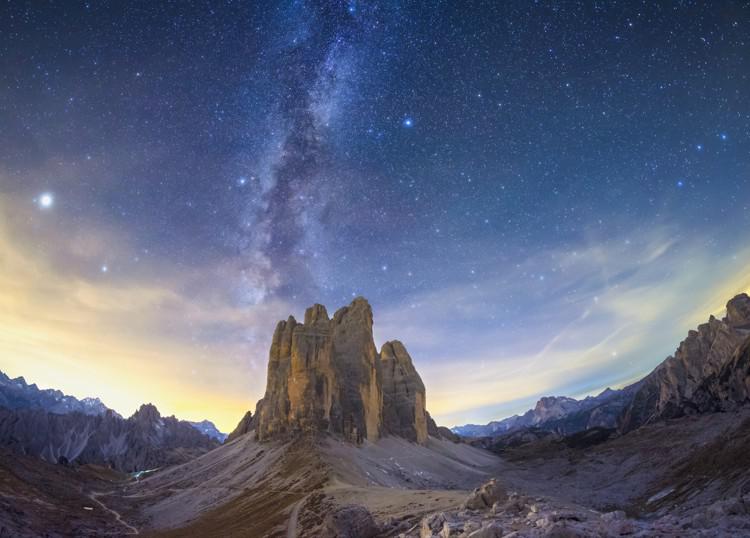 The Milky Way appears as two vertical diffuse bands of light either side of a dark line, over a rocky outcrop.