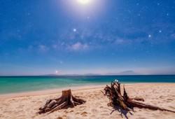 Der helle Mond beleuchtet einen Strand. Drei helle Planeten bilden eine Linie zum Horizont links unterhalb des Mondes.