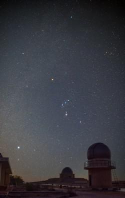 Orion appears as an hourglass with its belt slightly tilted relative to the horizon. The bright star Sirius is bottom-left