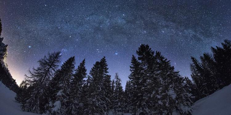 The bright, diffuse Milky Way, interrupted by mottled dark patches, arches over a wintry landscape.